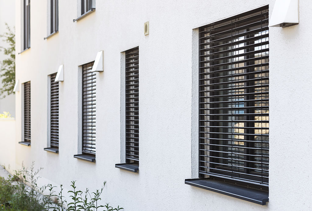 Mehrere Fenster mit geschlossenen Außenjalousien an der Fassade eines modernen Gebäudes.